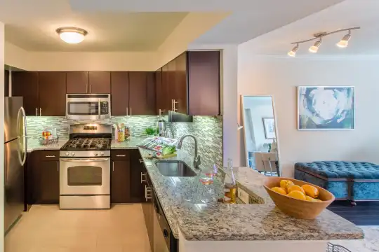 kitchen featuring stainless steel microwave, refrigerator, dishwasher, gas range oven, dark brown cabinetry, light tile flooring, and light stone countertops