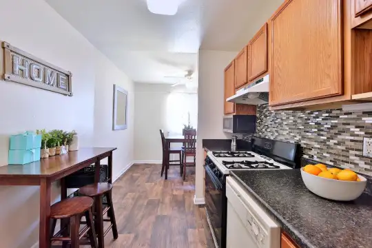 kitchen featuring range hood, gas range oven, dishwasher, dark granite-like countertops, brown cabinets, and light hardwood flooring